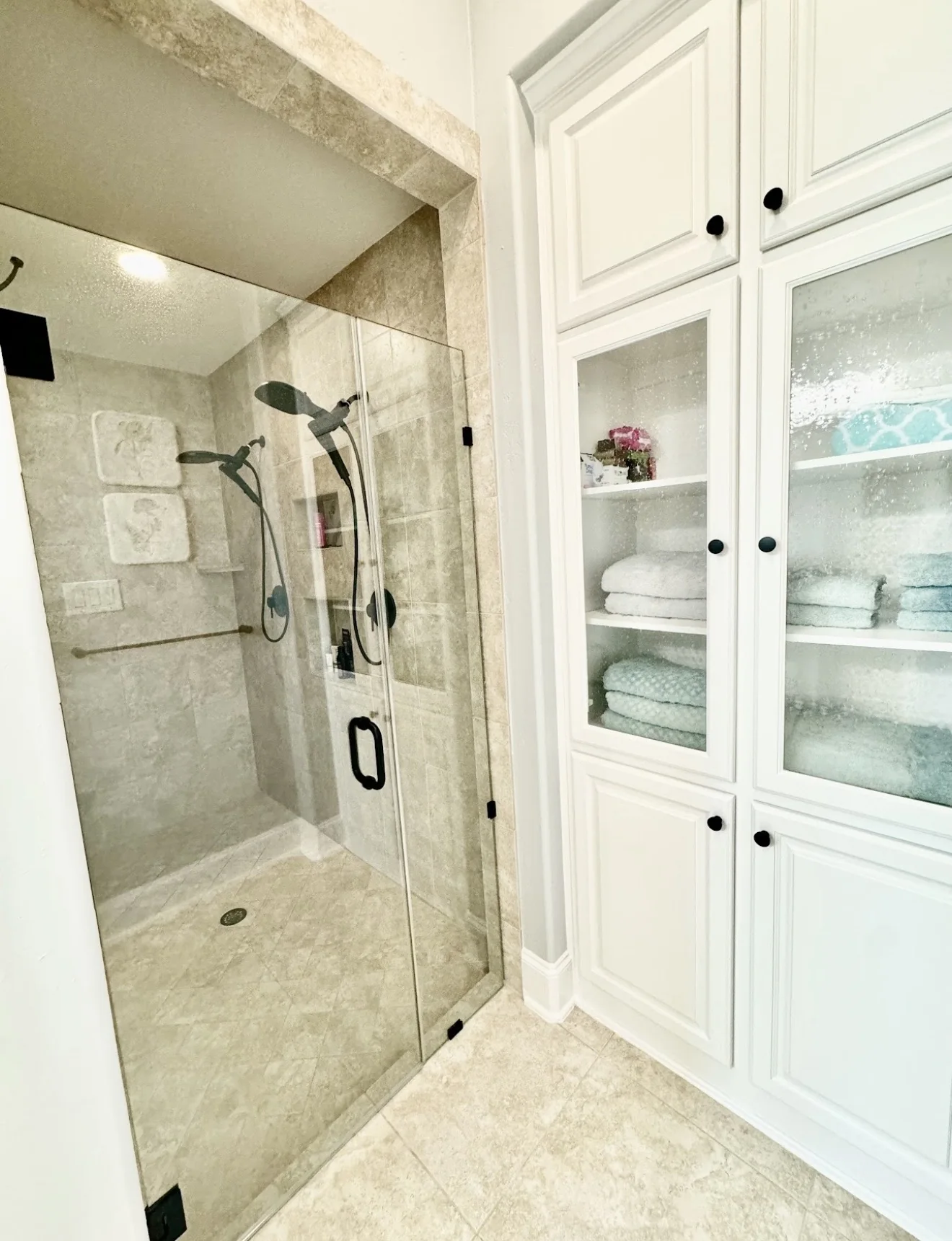 Custom walk-in shower with black fixtures and adjacent white linen cabinetry with glass doors