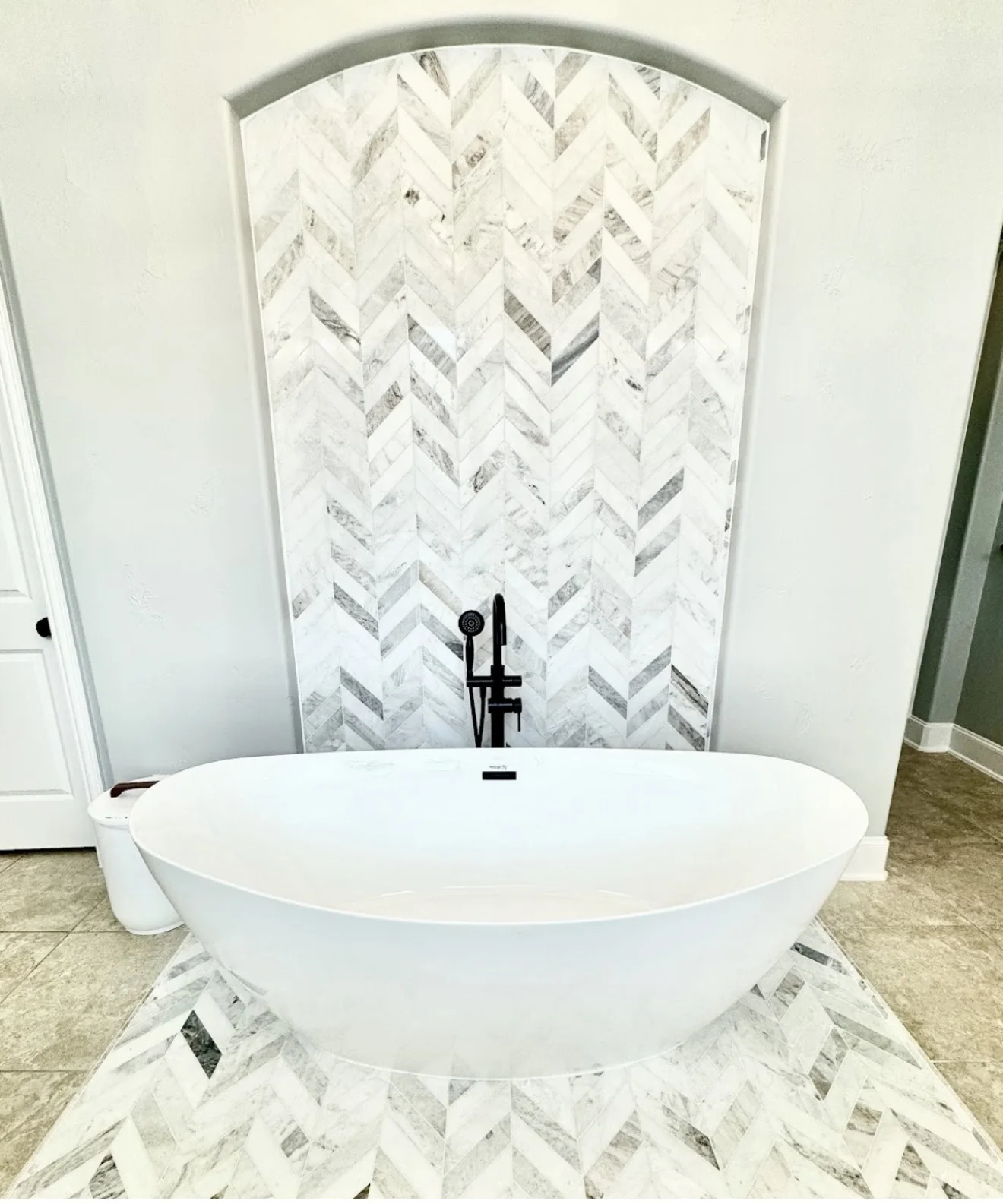 Freestanding soaking tub with marble-look herringbone accent wall and matte black floor-mounted tub filler